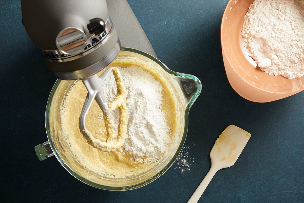 Overhead shot of cake batter being mixed in a stand mixer bowl, dry ingredients in a terracotta bowl beside it.