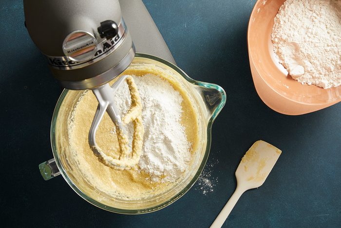 Overhead shot of cake batter being mixed in a stand mixer bowl, dry ingredients in a terracotta bowl beside it.