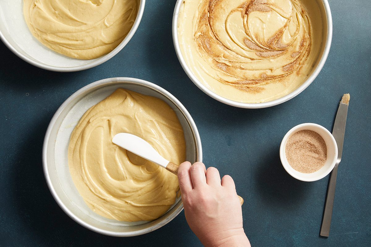 Overhead shot of two bowls of cake batter, one being smoothed with a spatula, set against a blue tabletop.