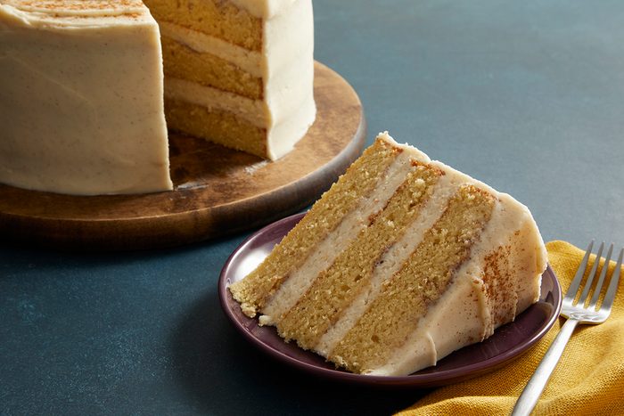 Close up shot of a slice of Snickerdoodle Cake on a plate, full cake on the stand in the background with a yellow napkin.
