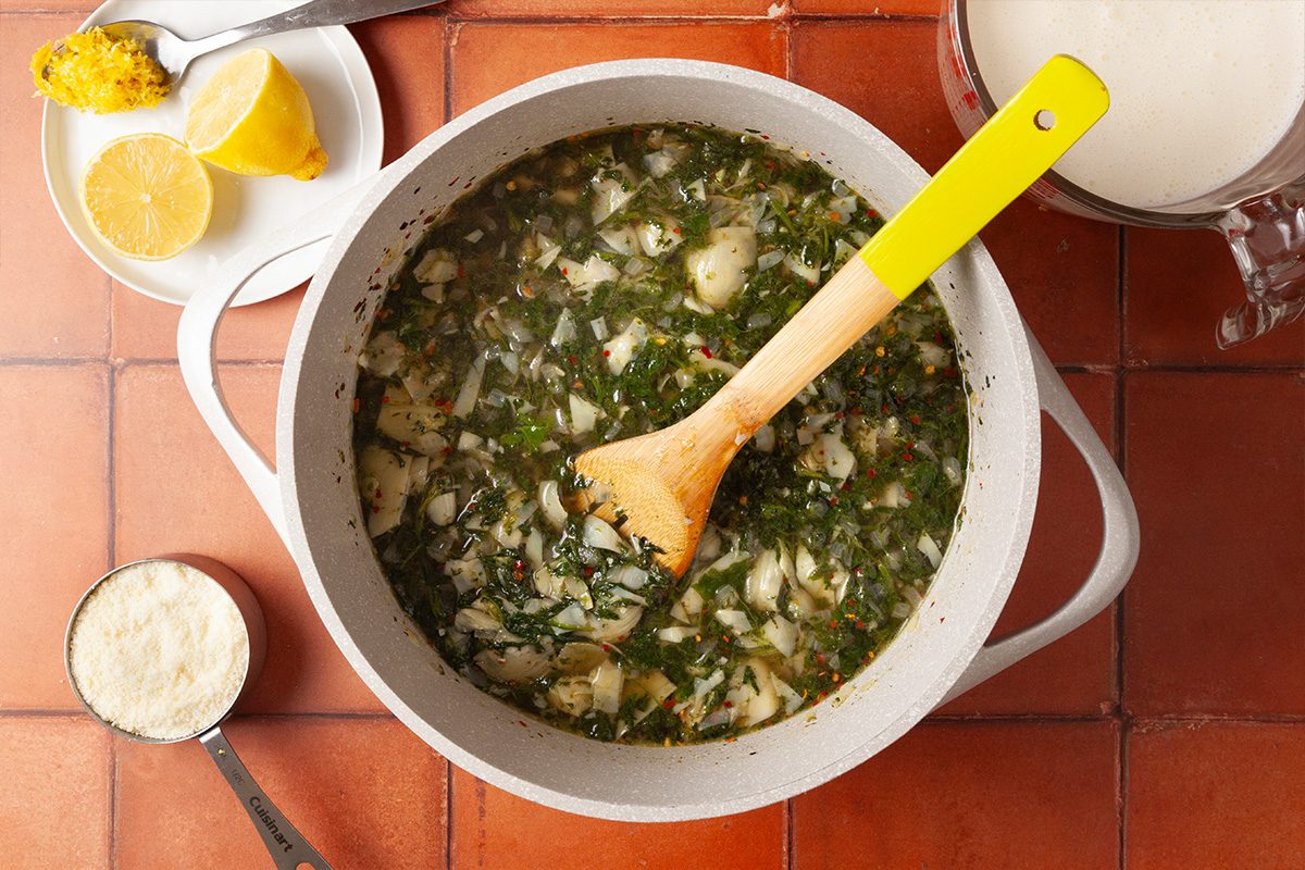 Overhead shot of a pot of soup with chopped greens and potatoes on a tiled surface, with a wooden spoon inside; Nearby are a cup of grated cheese, a cut lemon on a plate, and a pitcher of cream;