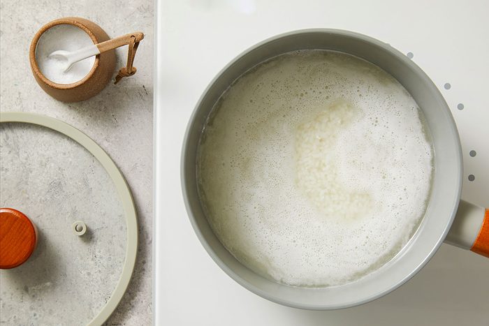 Overhead shot of a saucepan filled with rice simmering in boiling water on a stovetop; a small brown salt container with a white spoon sits nearby; part of a pot lid is visible on the countertop;