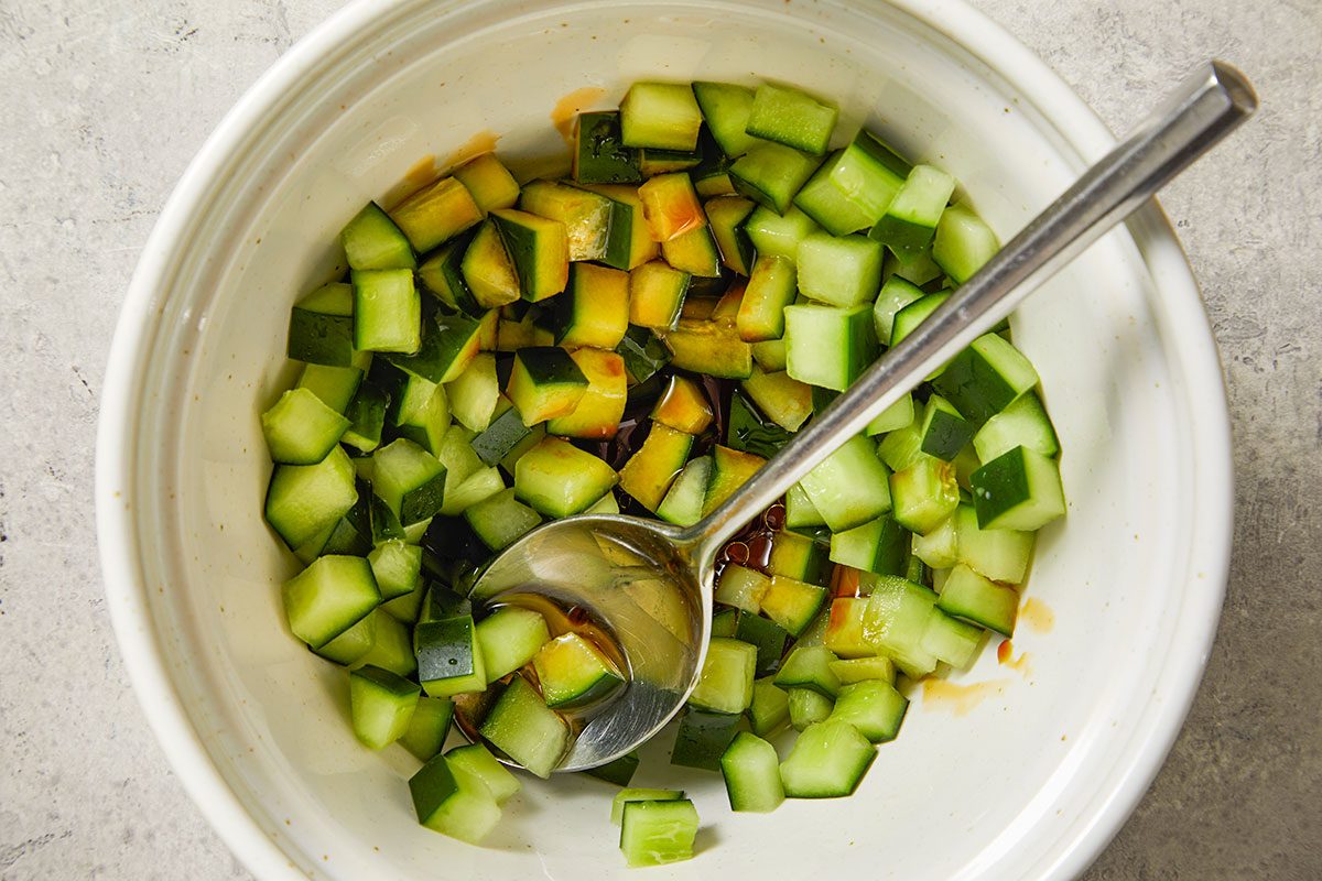Overhead shot of a white bowl filled with diced cucumber pieces; a metal spoon rests inside; a light brown dressing pools at the bottom; the bowl sits on a light-colored surface;