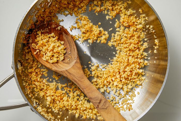 Overhead shot of a stainless steel pan filled with golden-brown breadcrumbs; a wooden spoon rests inside; toasted crumbs are scattered across the bottom of the pan;