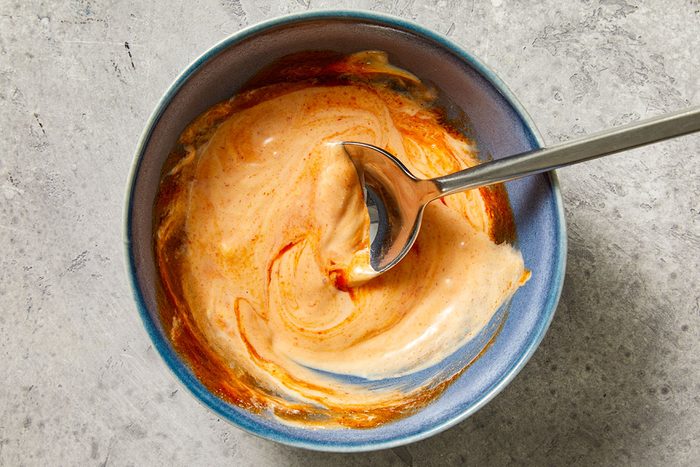 Overhead shot of a blue bowl filled with a creamy orange-and-white sauce; a silver spoon is stirring the mixture; set on a textured gray surface;
