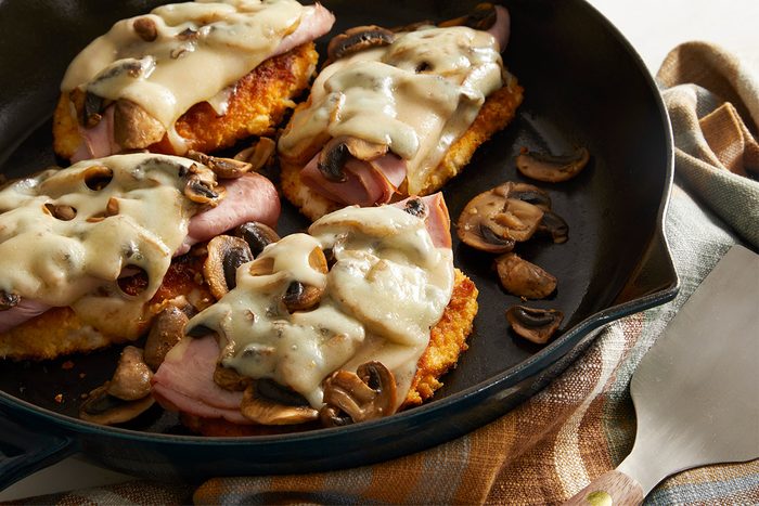 Close up shot of Swiss Mushroom Chicken topped with sautéed mushrooms, ham, and melted Swiss cheese in a black skillet on a plaid towel.