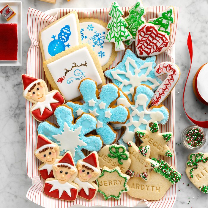 A tray of decorated holiday cookies, including snowflakes, elves, trees, candy canes, and gift tags with names. The cookies are colorful with red, green, blue, and white icing. A red ribbon and icing bowl are nearby.