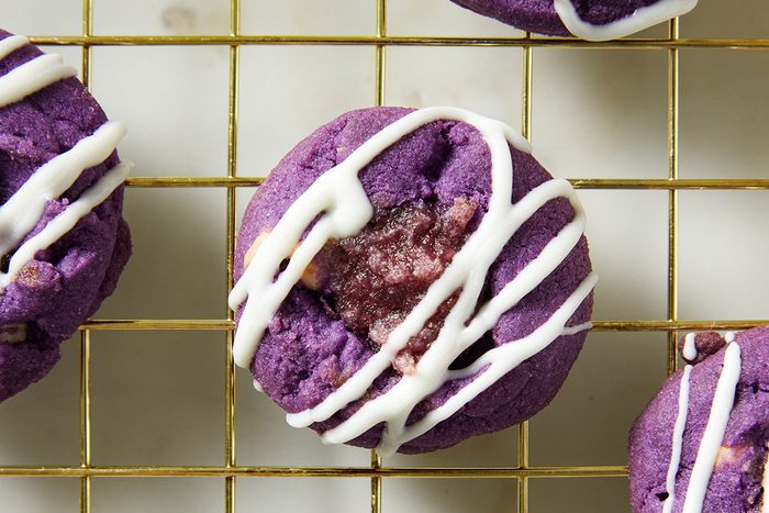 A vibrant purple cookie with a gooey center and drizzled with white icing, resting on a gold wire cooling rack.