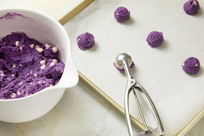 A baking sheet with scoops of purple cookie dough, some placed on parchment paper, sits next to a bowl of the same dough with white chocolate chips and a metal scoop.