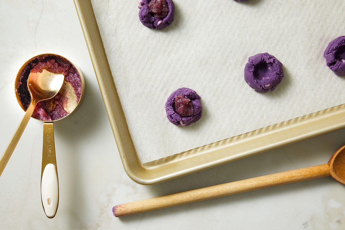 A baking tray with parchment paper holds several unbaked purple cookies. Next to it are a wooden spoon and a gold measuring cup with cookie dough on a white surface.
