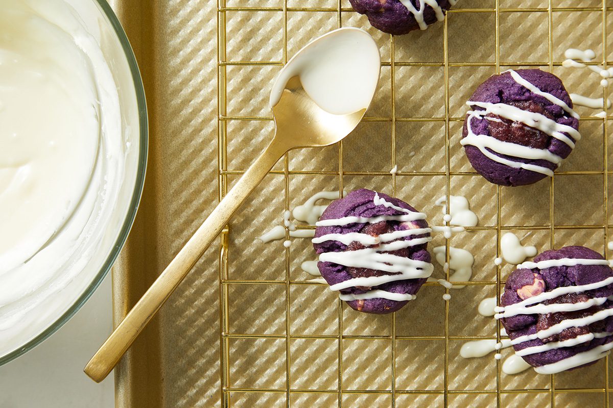 A gold spoon with white icing rests on a baking rack next to four purple cookies drizzled with icing. A glass bowl filled with more white icing sits nearby on the left.