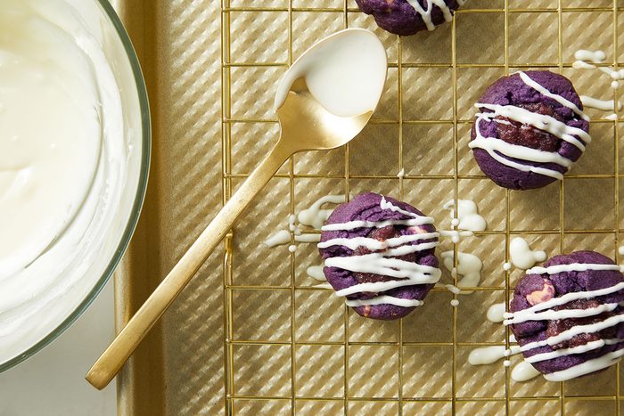 A gold spoon with white icing rests on a baking rack next to four purple cookies drizzled with icing. A glass bowl filled with more white icing sits nearby on the left.