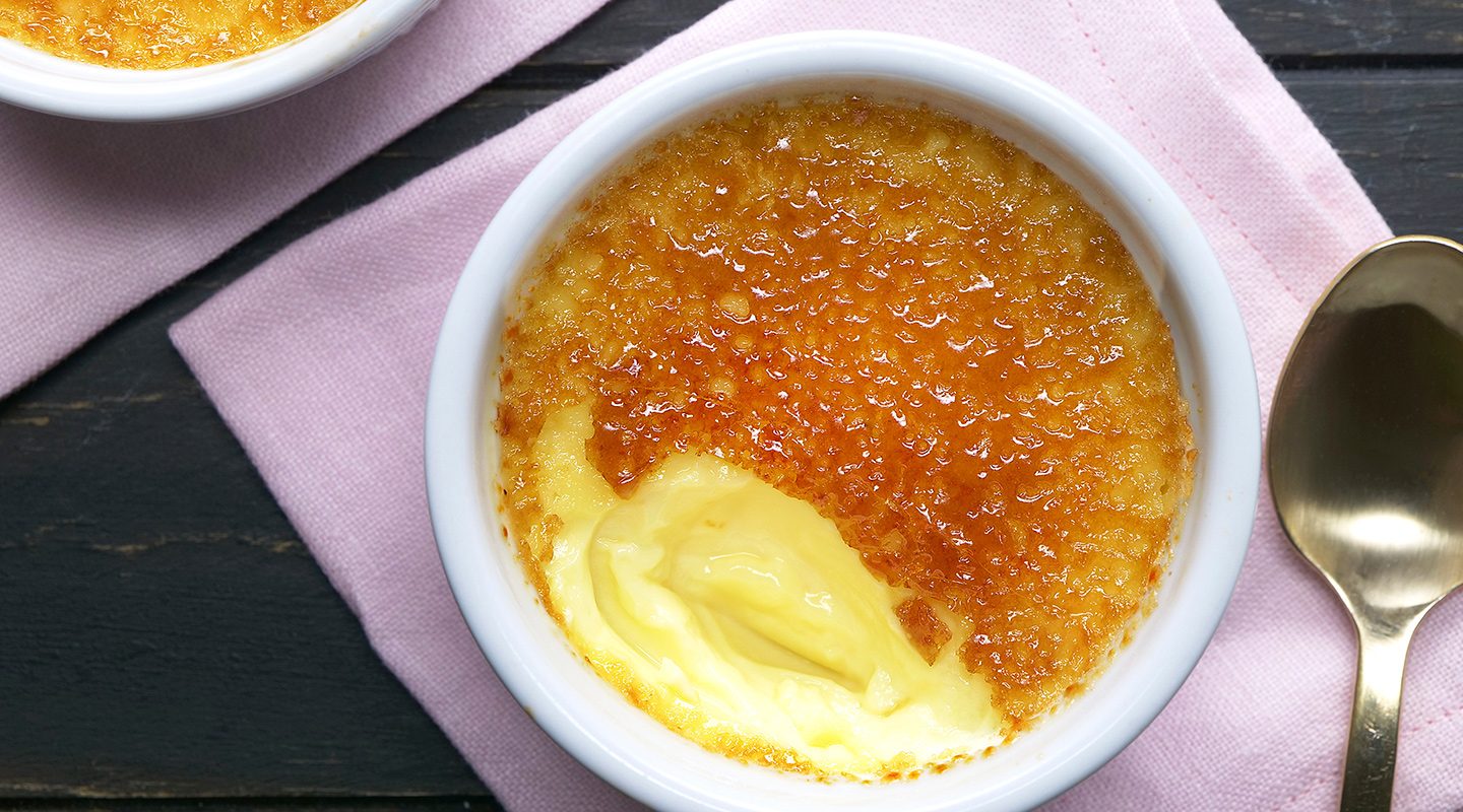 A ramekin of crème brûlée with a caramelized sugar crust, partially cracked to reveal creamy custard underneath, sits on a pink napkin next to a gold spoon.