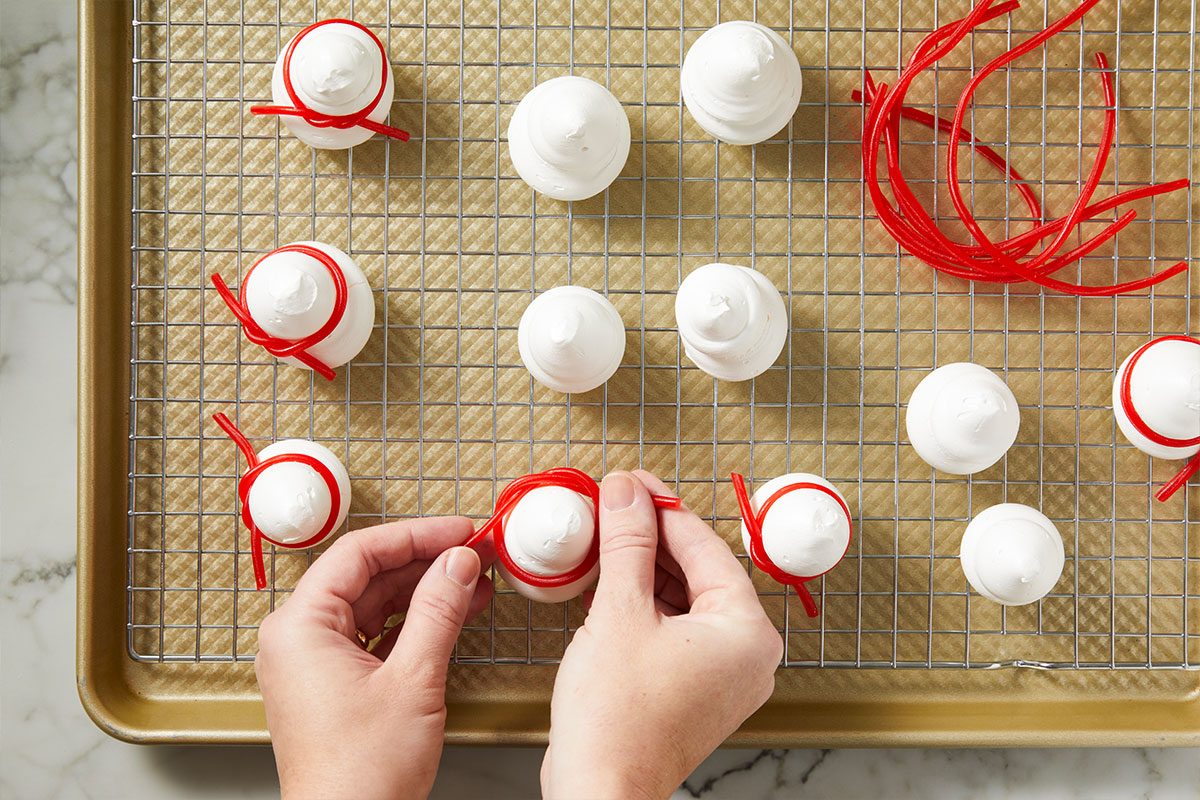 Overhead shot of Hands are tying red candy rope on meringue cookies shaped like hats as they sit on a rack over parchment paper and some are decorated