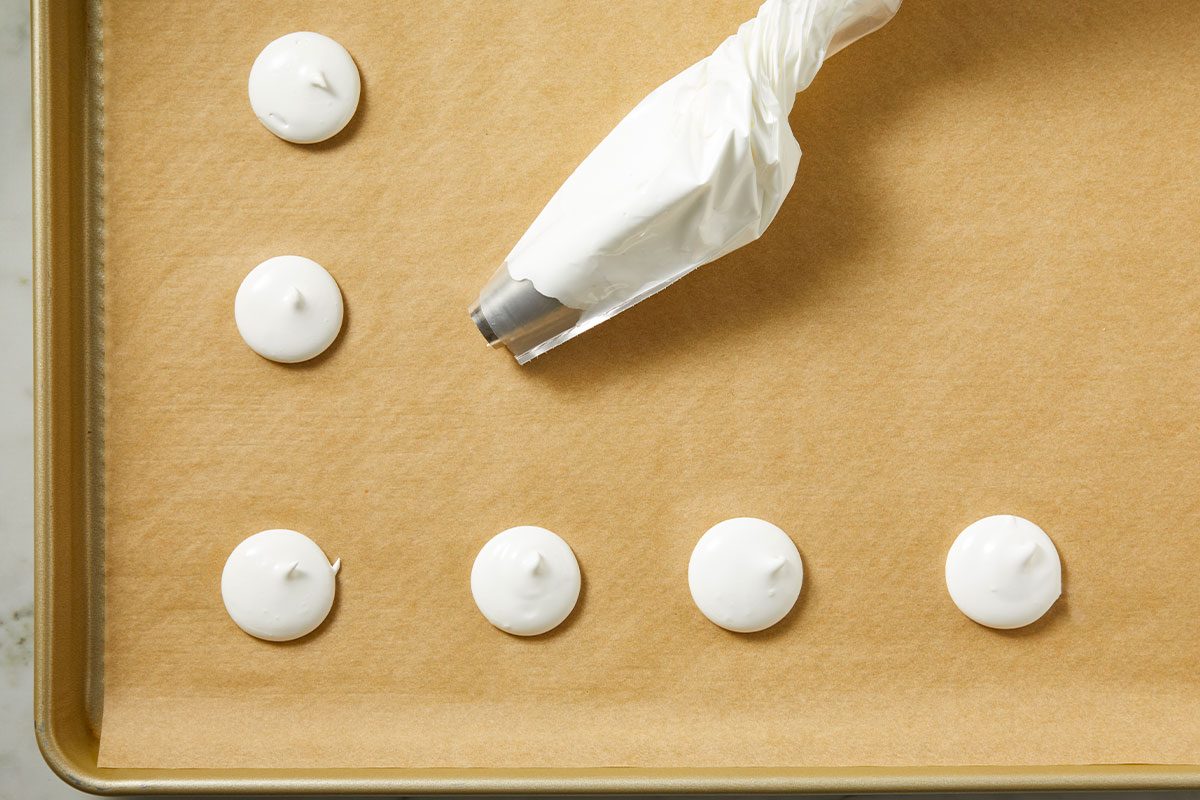 Overhead shot of a piping bag squeezes out white meringue onto a parchment-lined baking tray neat round dollops appear in a single row