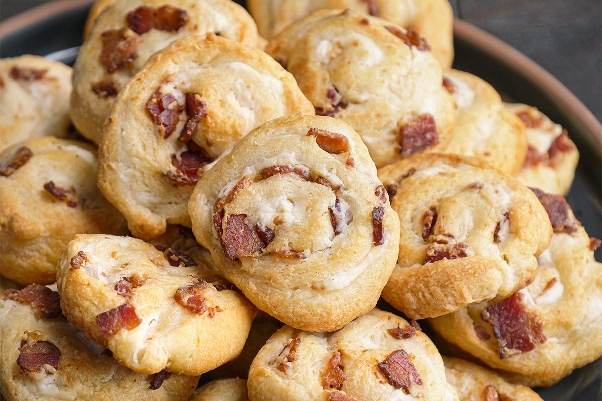This is a close-up shot of a plate stacked with golden-brown pinwheel pastries; bacon; cheese fillings; white background