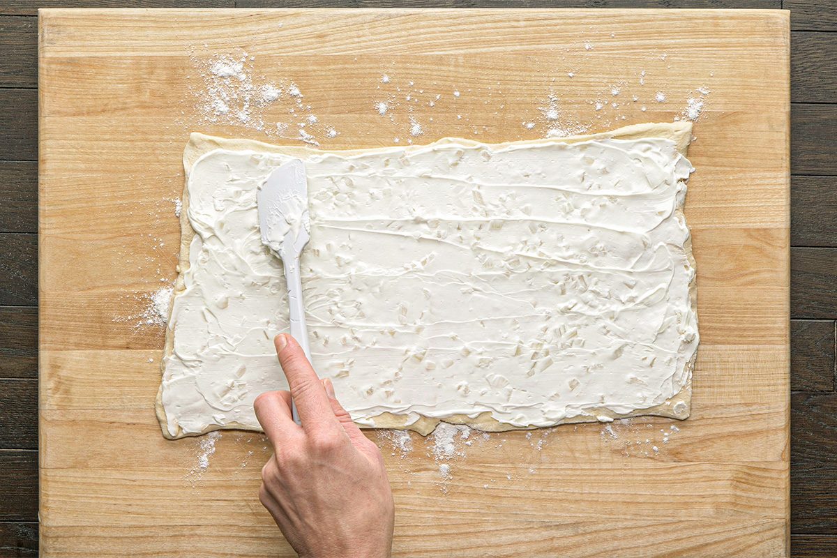 This is a close-up shot; a hand uses a white spatula to spread cream cheese on rectangular dough; floured wood background