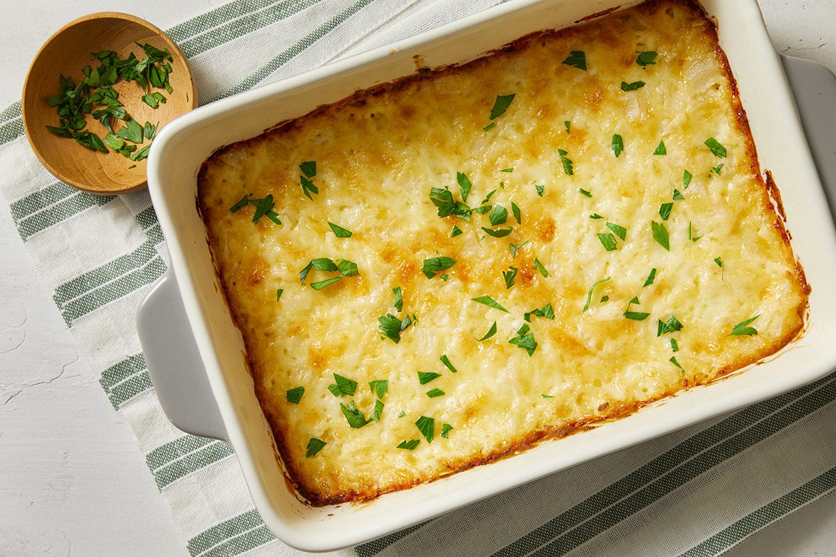 A baked casserole topped with melted cheese and sprinkled with fresh herbs, in a white rectangular dish on a striped cloth, with a small bowl of chopped herbs nearby.