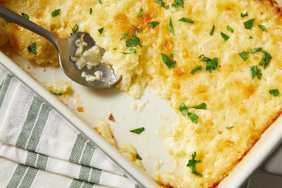 A baking dish of creamy, golden-brown casserole topped with melted cheese and chopped parsley. A serving spoon rests in the dish, with a portion already scooped out. A striped cloth is nearby.