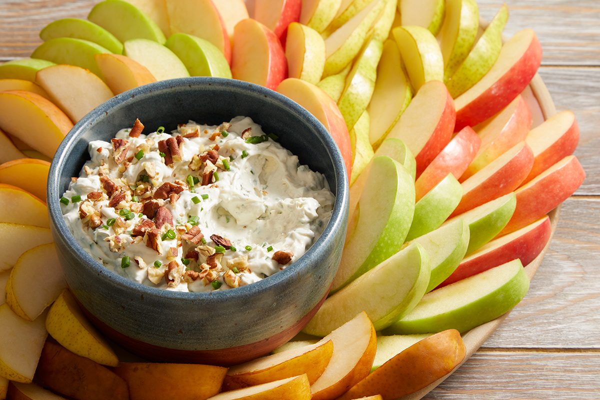 A bowl of creamy dip garnished with chopped nuts and herbs is surrounded by neatly arranged slices of red, green, and yellow apples on a wooden platter.
