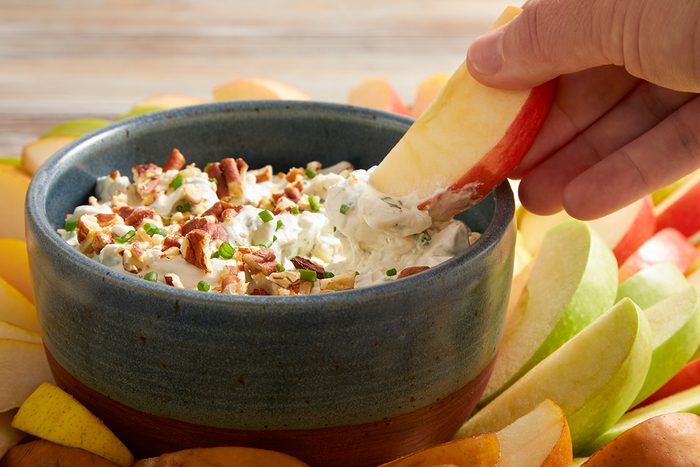 A hand dips an apple slice into a bowl of creamy dip topped with chopped pecans and chives, surrounded by assorted apple slices.