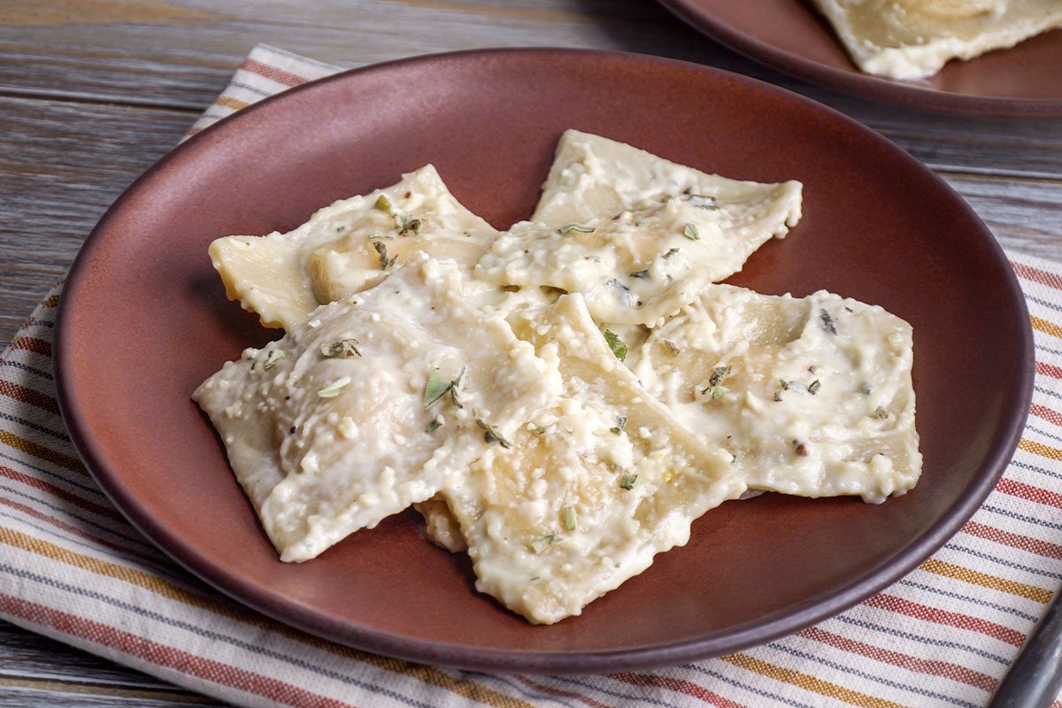 3/4 angle view shot of Butternut Squash Ravioli and it is served on a brown plate with creamy white sauce and fresh herbs on top and the plate sits on a striped napkin placed on a wooden table