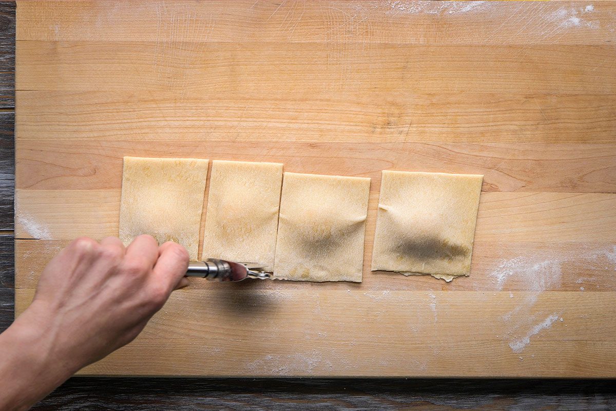 Overhead shot of a hand uses a fork to press and seal the edges of four rectangular pieces of dough on a floured wooden surface sealing each piece