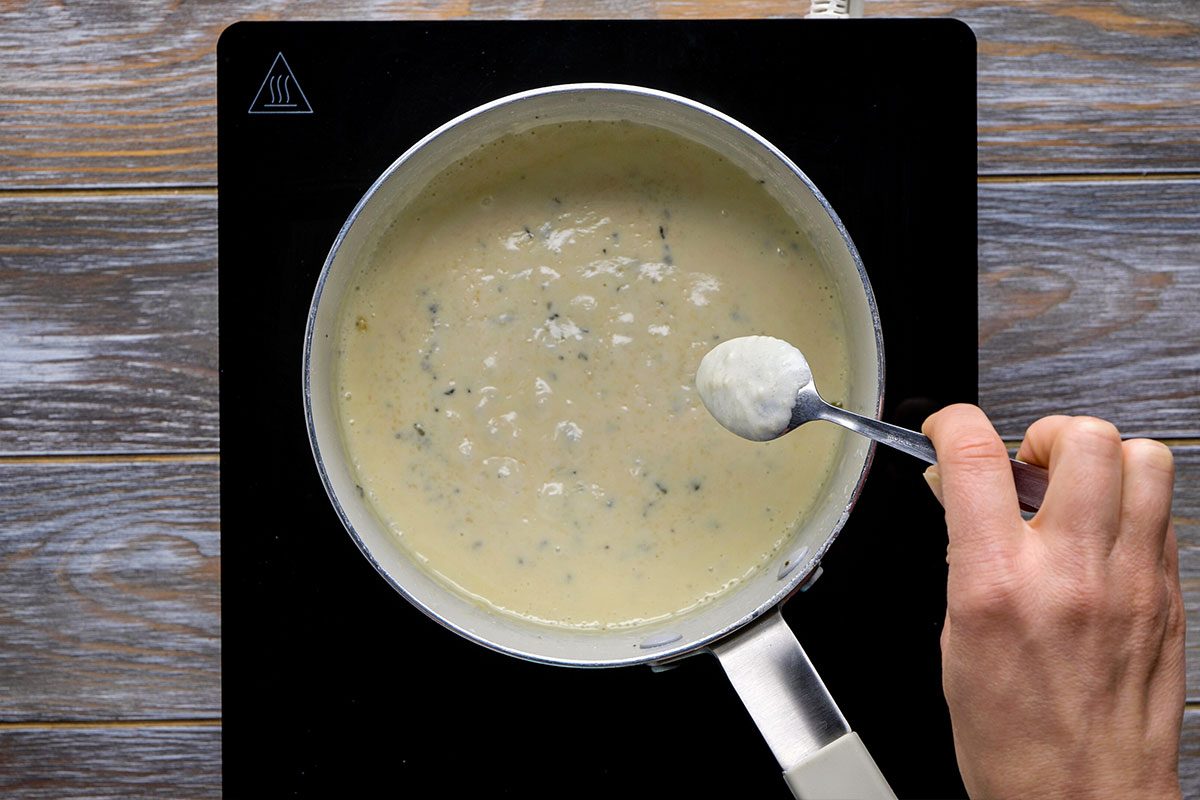 Overhead shot of a hand holds a spoon over a saucepan filled with creamy white herb sauce it is heating on a black stovetop placed on wooden surface