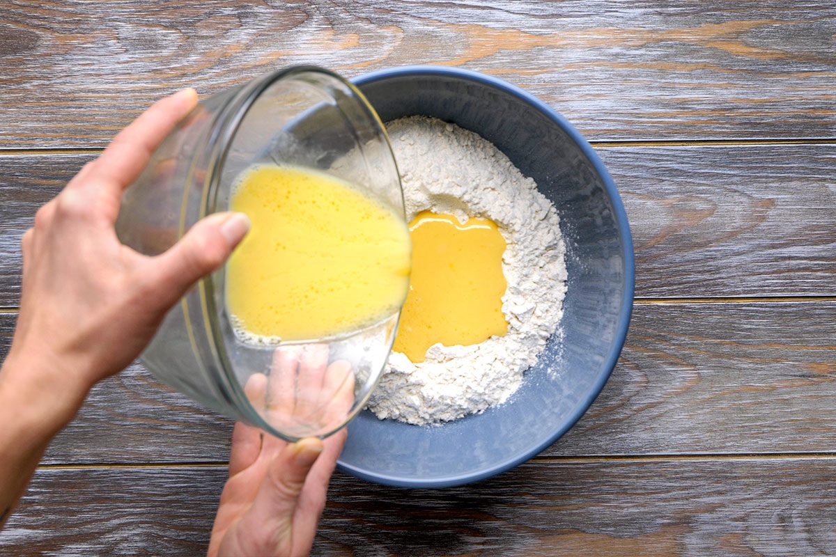 Overhead shot of a person begins to pour a bowl of yellow liquid into a larger blue bowl filled with flour on a wooden surface and readies the mix together