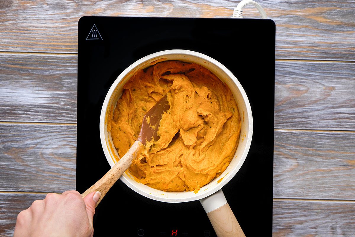 Overhead shot of a person stirs thick orange mashed butternut squash in a white saucepan using a black spatula and the pot sits on a dark potholder on a wooden surface