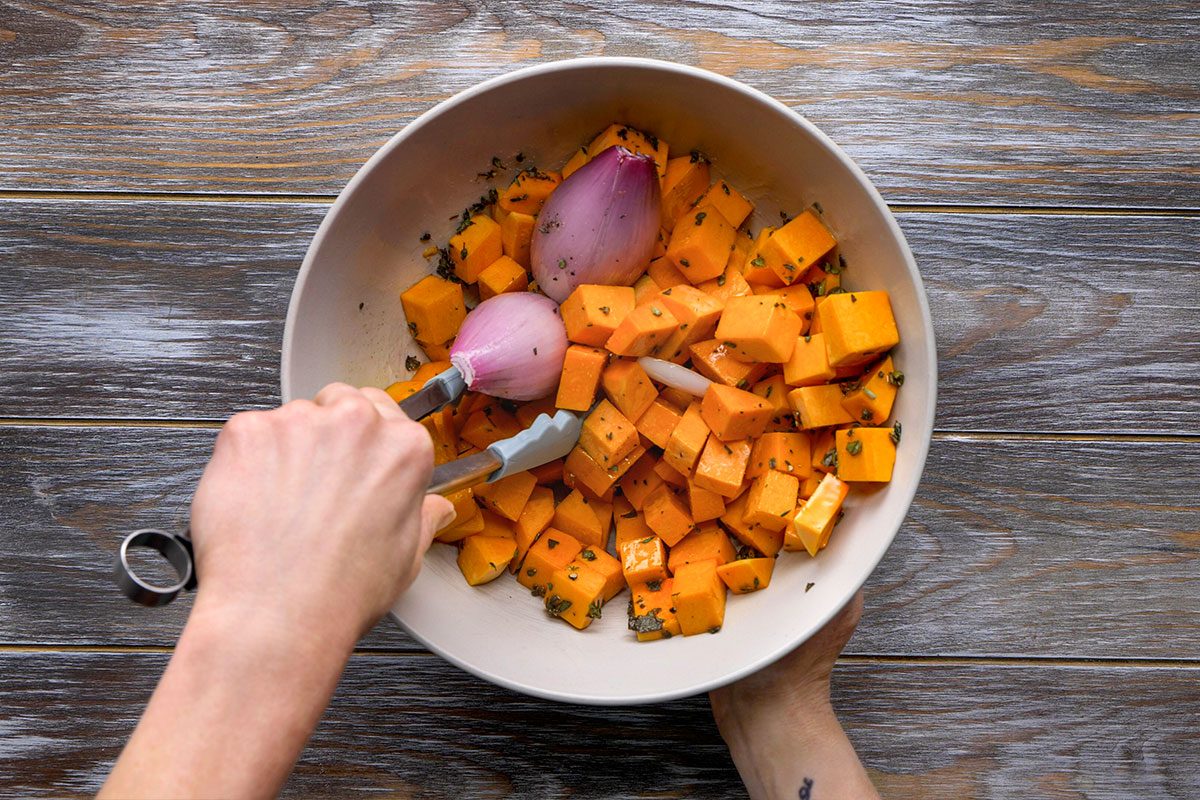 Overhead shot of a person uses tongs to mix cubed butternut squash and halved shallots with herbs in a big white bowl on a wooden surface The mix is fresh and colorful