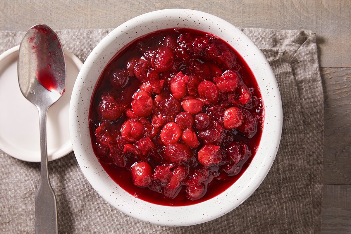 A white bowl filled with chunky cranberry sauce sits on a gray cloth napkin. A spoon with cranberry sauce rests on a small white plate next to the bowl.