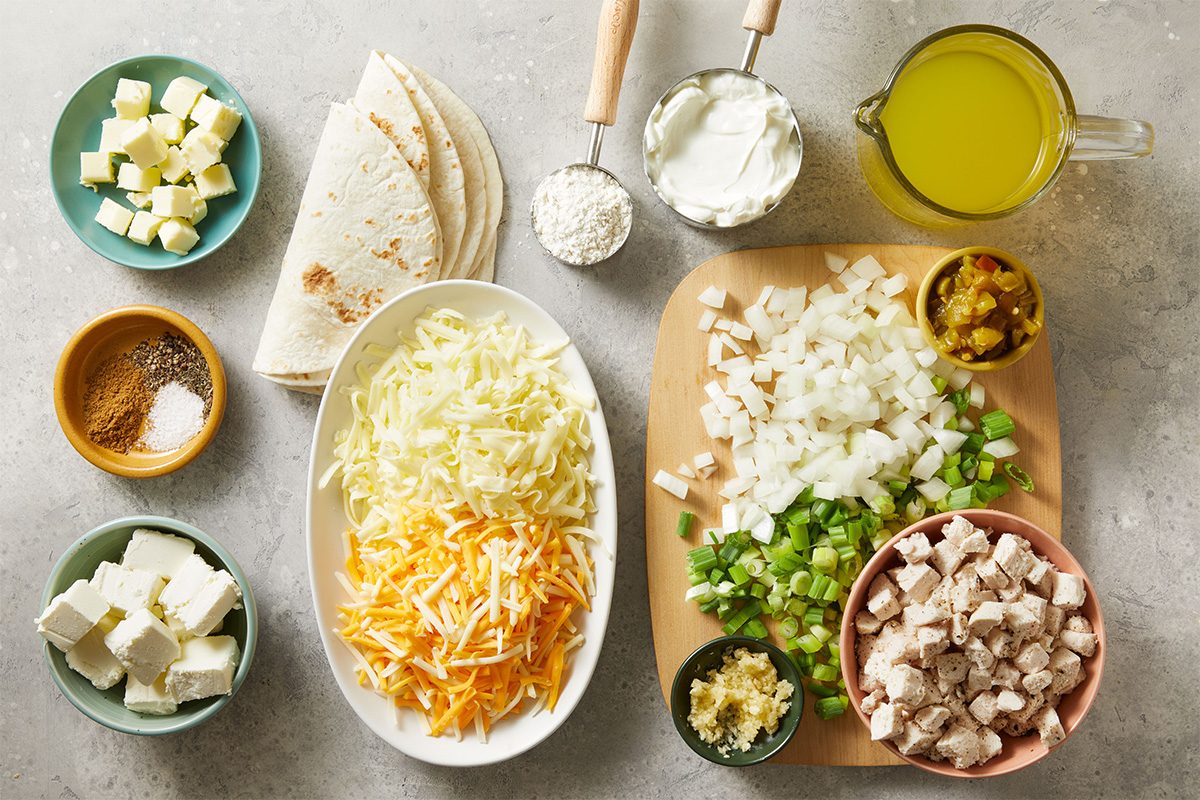 Overhead shot of neatly arranged recipe ingredients on a light surface, including shredded cheese, chopped onions, tortillas, cooked chicken, sour cream, green chiles, broth, and spices.