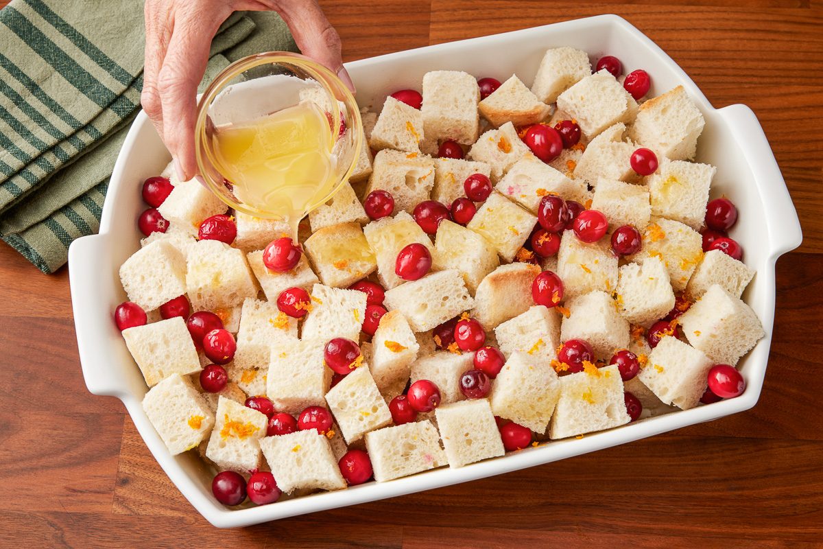 bread cubes, cranberries and orange zest layered in a greased 13x9-inch baking dish being drizzled with butter