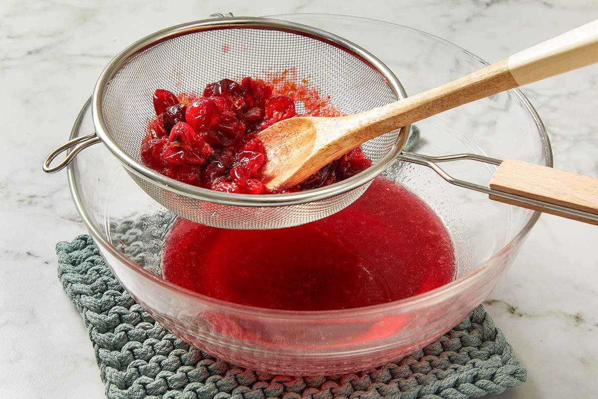 Overhead shot of cranberry mixture being strained through a fine-mesh sieve into a glass bowl, wooden spoon pressing the fruit to release bright red syrup.