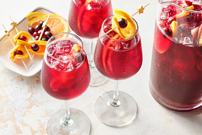 3/4 shot of several glasses of Cranberry Orange Sangria on a light surface, each topped with cranberries and orange slices, with a pitcher beside them.