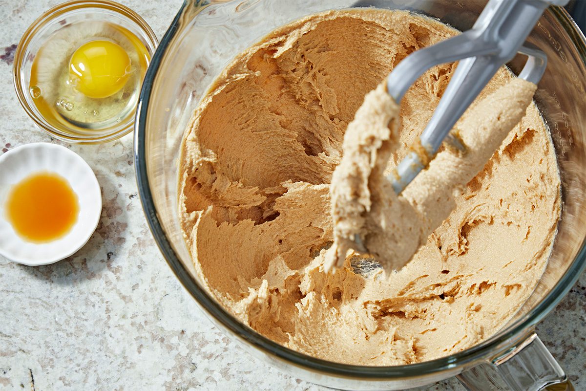 A mixing bowl with creamy cookie dough and a paddle attachment, next to a small bowl with a cracked egg and another small bowl with vanilla extract, all on a light countertop.