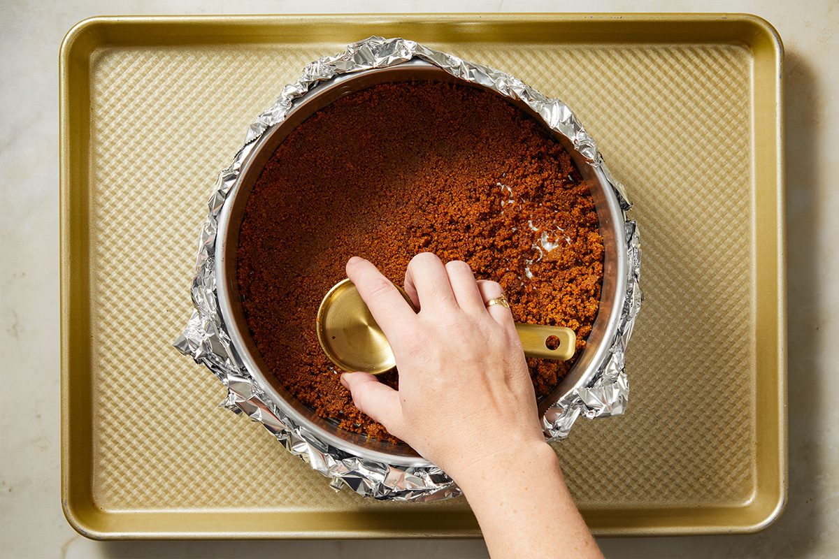 Overhead shot of graham cracker crumbs being pressed firmly into the bottom of the springform pan using a flat measuring cup to form an even crust.