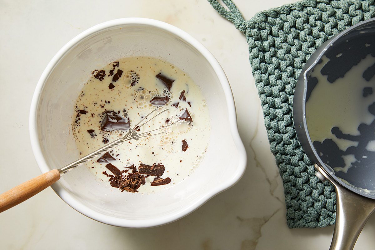 Overhead shot of warm cream in a bowl with chopped chocolate melting into it, being stirred into a smooth ganache.