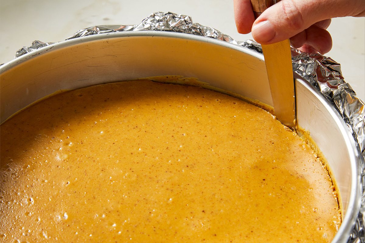 Overhead shot of the baked cheesecake with a golden surface, a knife running around the edge to loosen it from the sides of the pan.