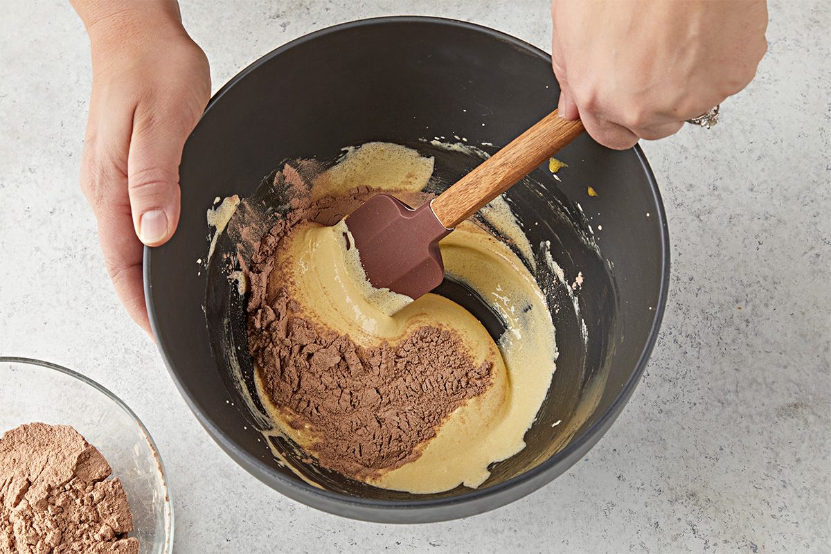 Overhead shot of a person using a spatula to mix cocoa powder into a bowl of yellow batter, preparing a chocolate mixture on a light kitchen countertop;