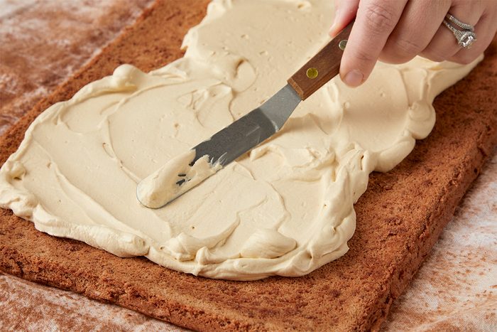 Closeup shot of a hand using a spatula to spread a thick layer of creamy frosting over a rectangular sheet of cake on a brown surface;