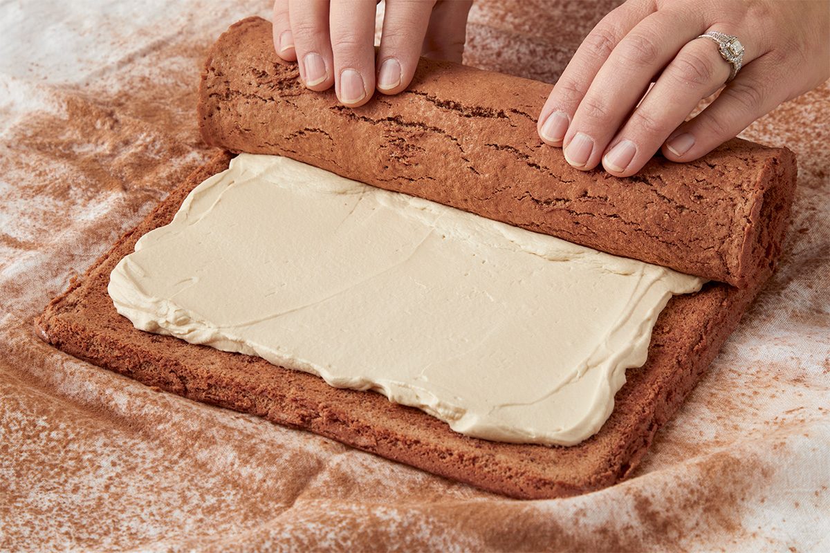 3/4 angle view shot of hands rolling up a rectangular sheet cake spread with a layer of cream filling on a lightly floured surface;