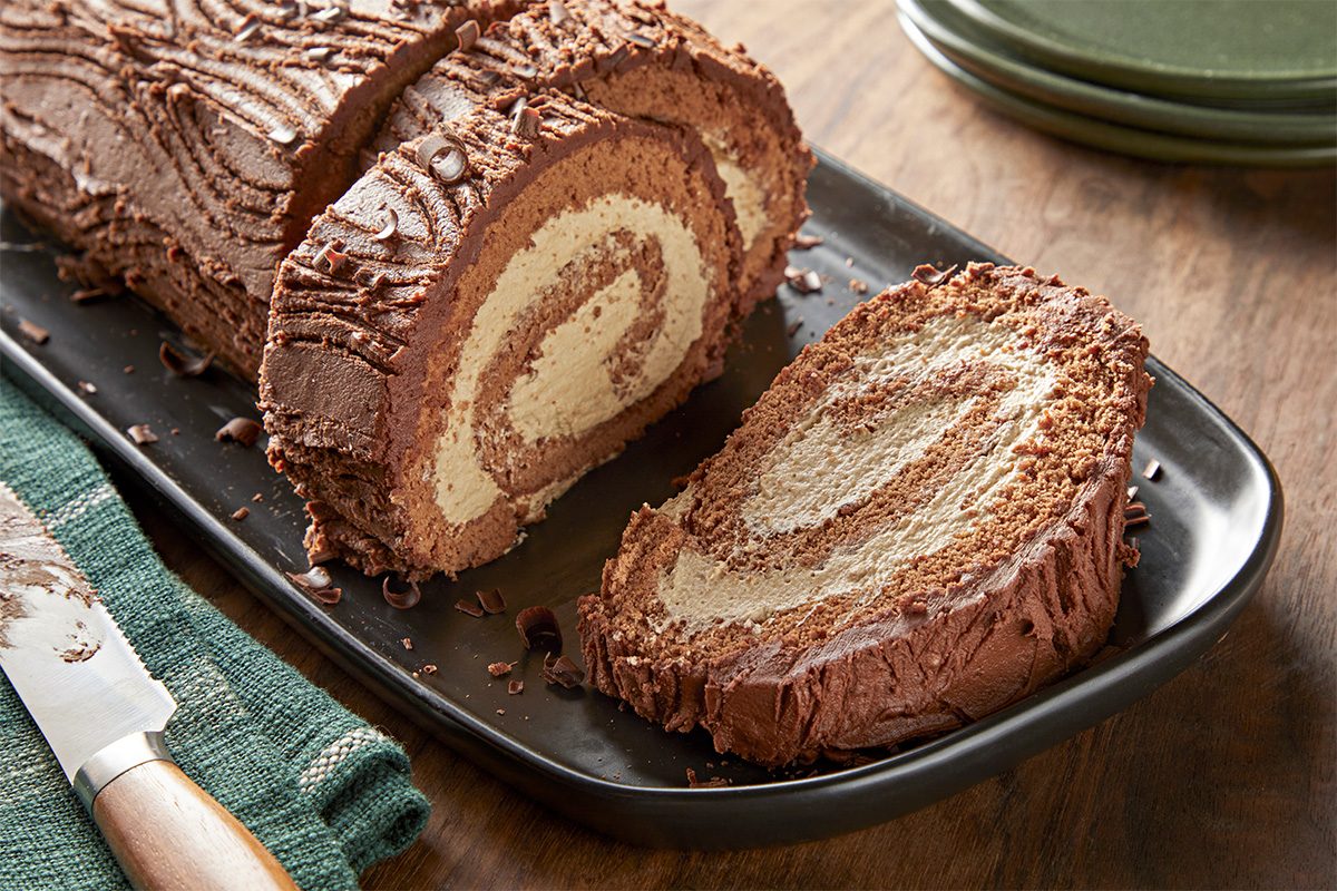 Close shot of a Mocha Yule Log with creamy swirl filling, partially sliced on a black platter on a wooden table, beside a knife and green napkin;