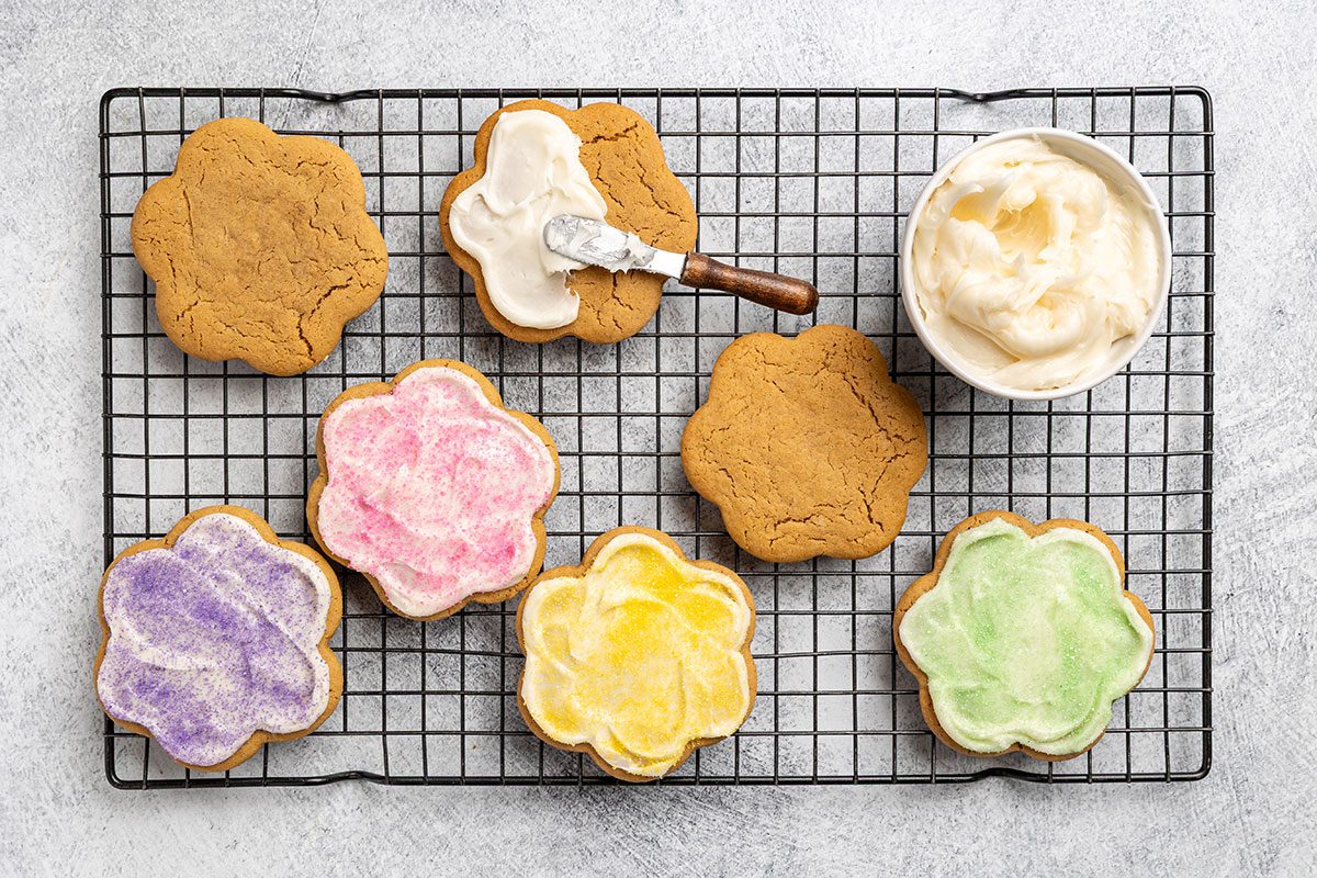Overhead shot of flower-shaped cookies on a cooling rack; some unfrosted and others topped with pastel-colored frosting; with a knife and a bowl of white frosting also on the rack.