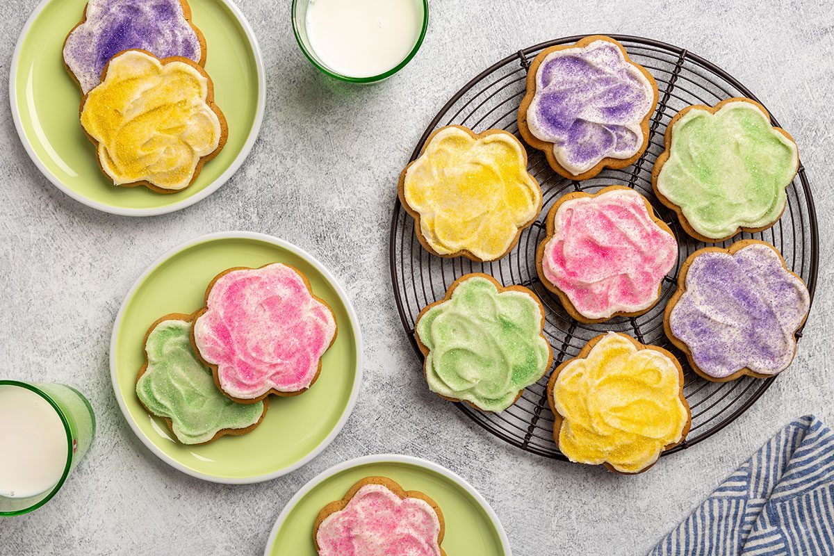 Overhead shot of Peanut Butter Cutout Cookies shaped like flowers; decorated with pastel-colored frosting in pink, yellow, green, and purple; Some cookies are arranged on a cooling rack; while others are placed on green plates with glasses of milk nearby; all set on a light textured surface.