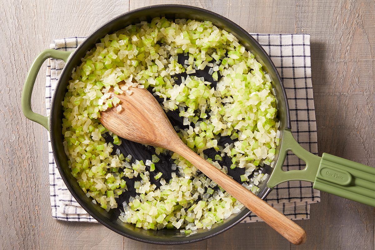 Overhead shot of chopped onions and celery sautéing in butter in a green skillet with a wooden spoon resting inside.