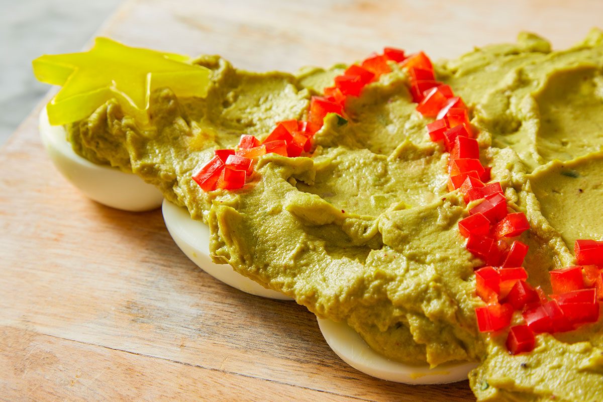 Overhead shot of a Pull-Apart Deviled Egg Christmas Tree is arranged on a Christmas tree-shaped platter topped with guacamole red pepper garland and a yellow star while three forks sit on a plaid napkin beside the wooden board