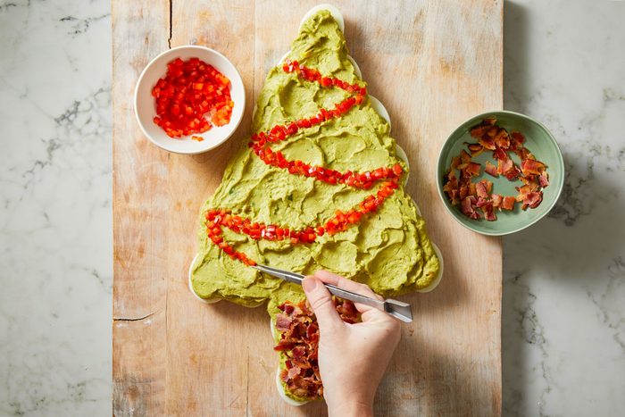 Overhead shot of a hand places diced red peppers and bacon on a Christmas tree-shaped spread set on a wooden board Bowls with extra toppings are nearby