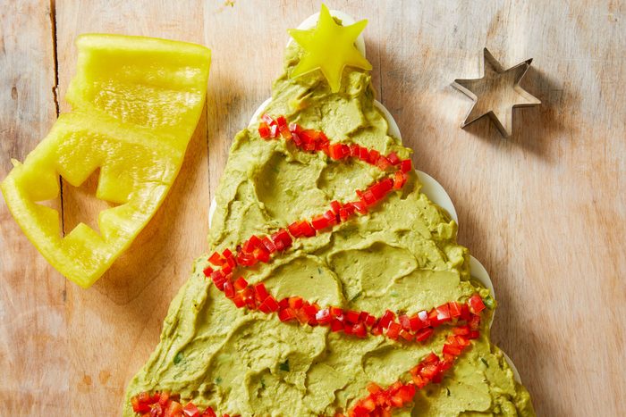 Overhead shot of a platter of guacamole shaped like a Christmas tree is decorated with red and yellow bell pepper A metal cookie cutter sits nearby